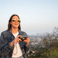 Woman wearing sunglasses, holding a camera in front a of cityscape down the mountains