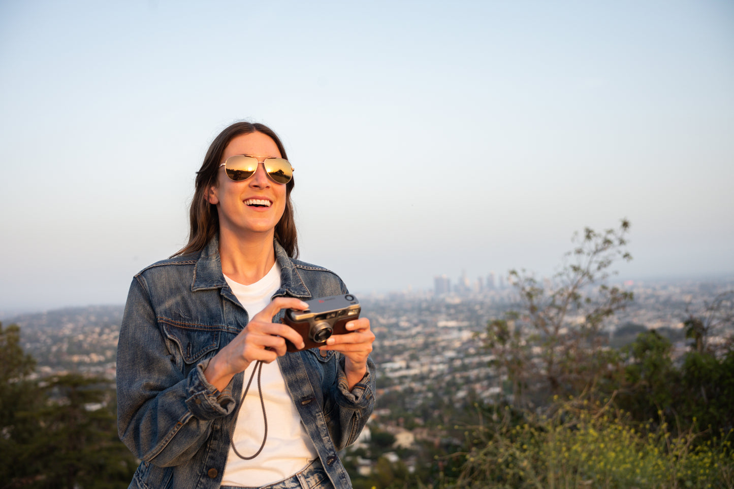 Woman wearing sunglasses, holding a camera in front a of cityscape down the mountains