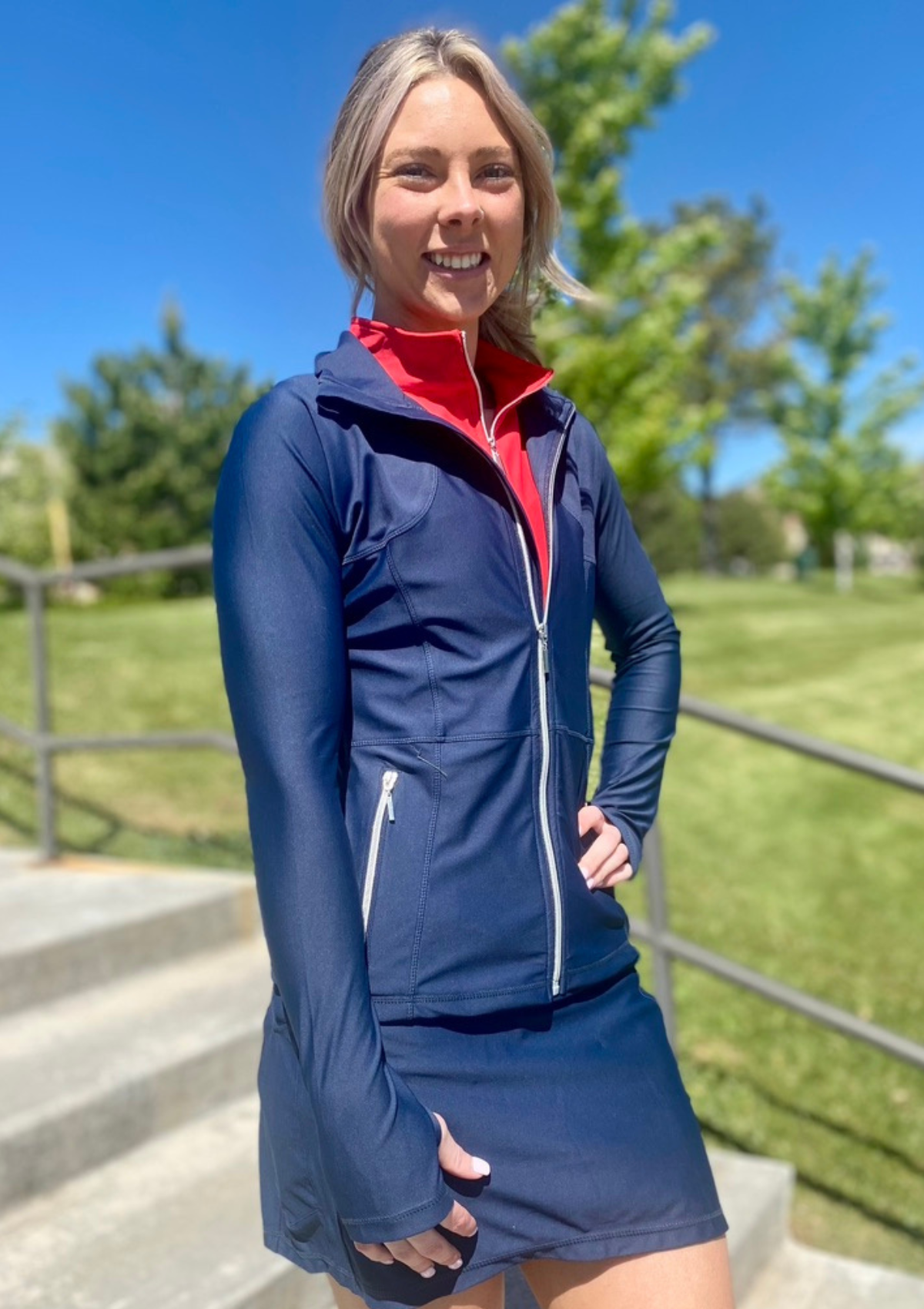 Woman outdoors on stairs in navy full-zip jacket, red top underneath and navy skort