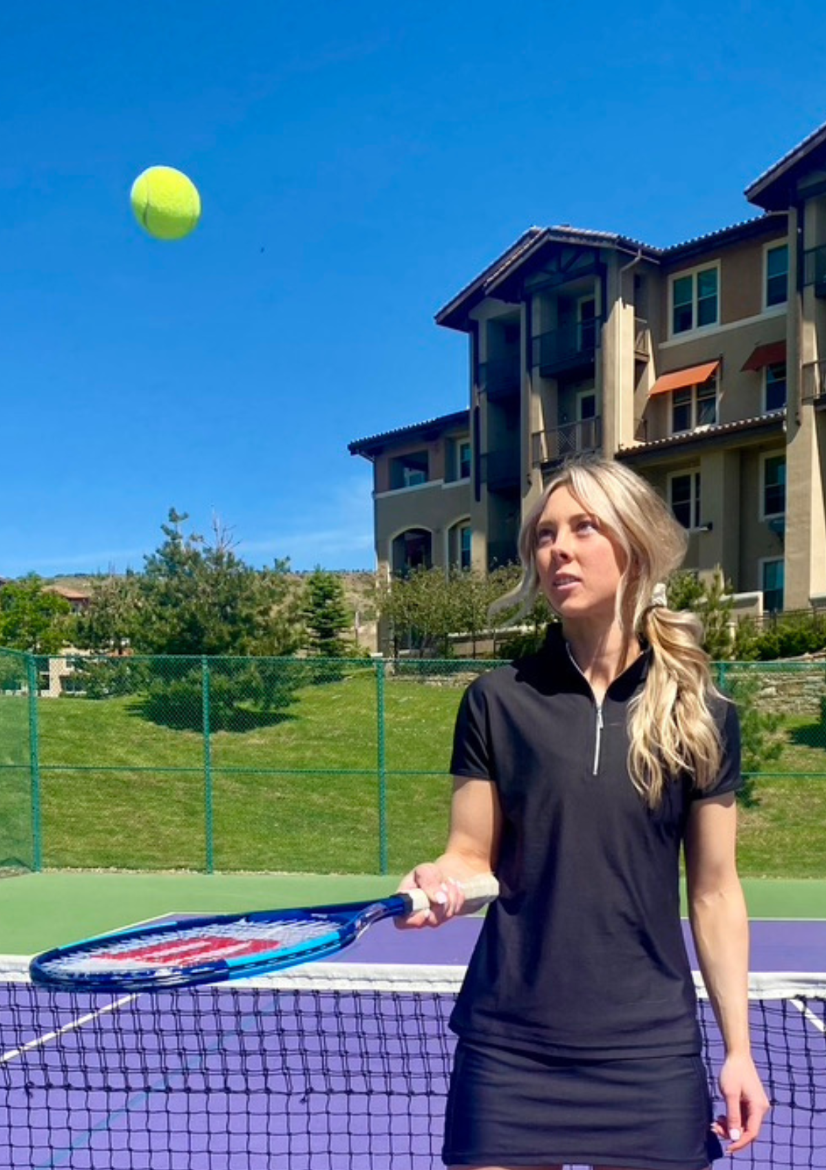 Woman bouncing tennis ball on court in front of mountainside home